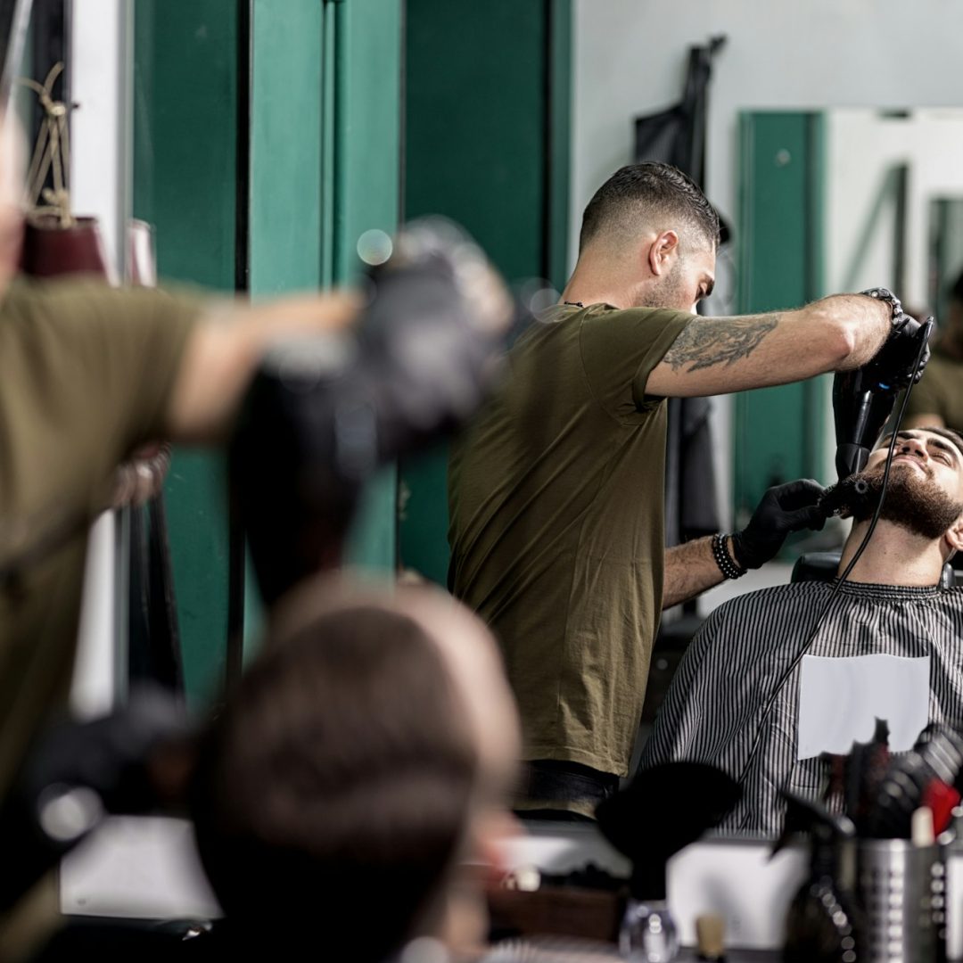 Brutal man with a beard sits in front of the mirror at a barbershop. Barber trims mens beard with