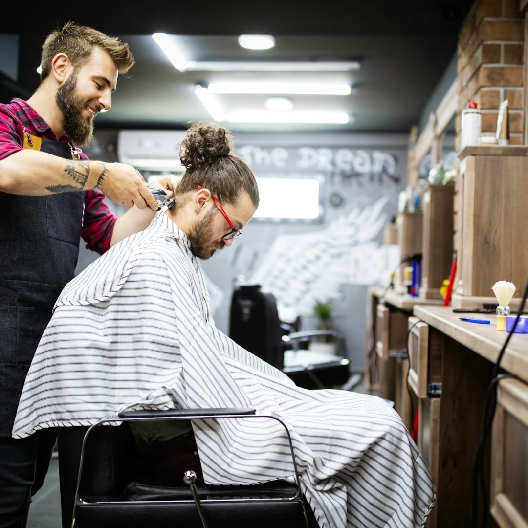 Young bearded man getting haircut by hairdresser while sitting in chair at barbershop